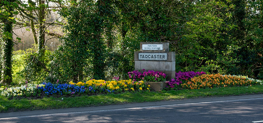 sign of tadcaster on a rock with flowers surrounding it on the side of the road